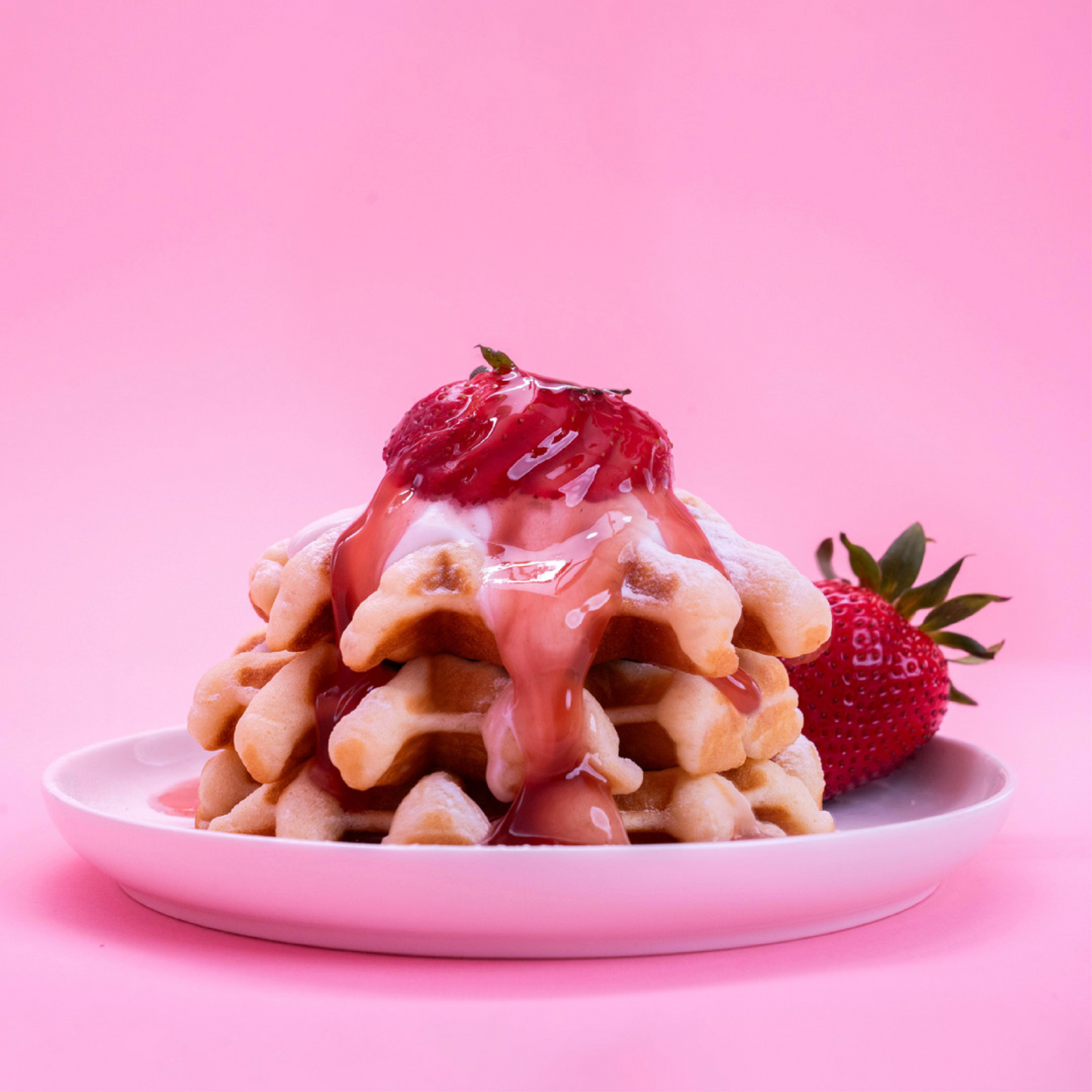 A stack of waffles topped with Lakanto Sugar-Free Strawberry Syrup and fresh berries sits on a white plate. The solid pink background enhances the vibrant scene, with a whole strawberry beside this keto-friendly delight.