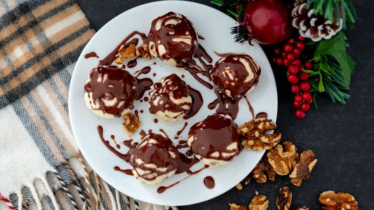 Six white maple walnut truffles are arranged in a star fashion on a white plate. Chocolate is drizzled heavily on them. The plates sits on the corner of a white, gold, and black cloth. Walnuts are scattered in the bottom right corner.