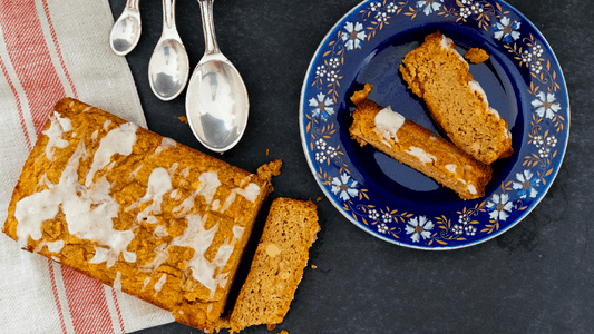 A loaf of low carb pumpkin bread is displayed on a dark tabletop in the bottom left corner. A piece is sliced off and lays in where it fell. The loaf is drizzled with white frosting. Two other pieces are on a blue, flowered plate in the top right corner. 