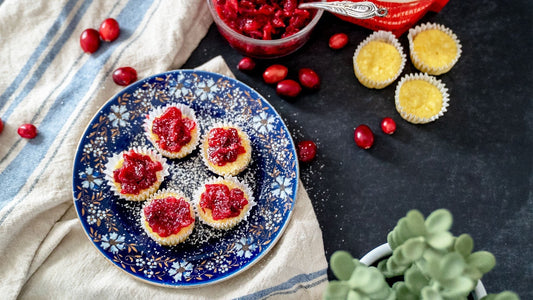 A blue plate with white and brown flowers sits on a white with blue lines dish towel. Cranberries, cupcakes, and Lakanto sweetener are scattered around the table.