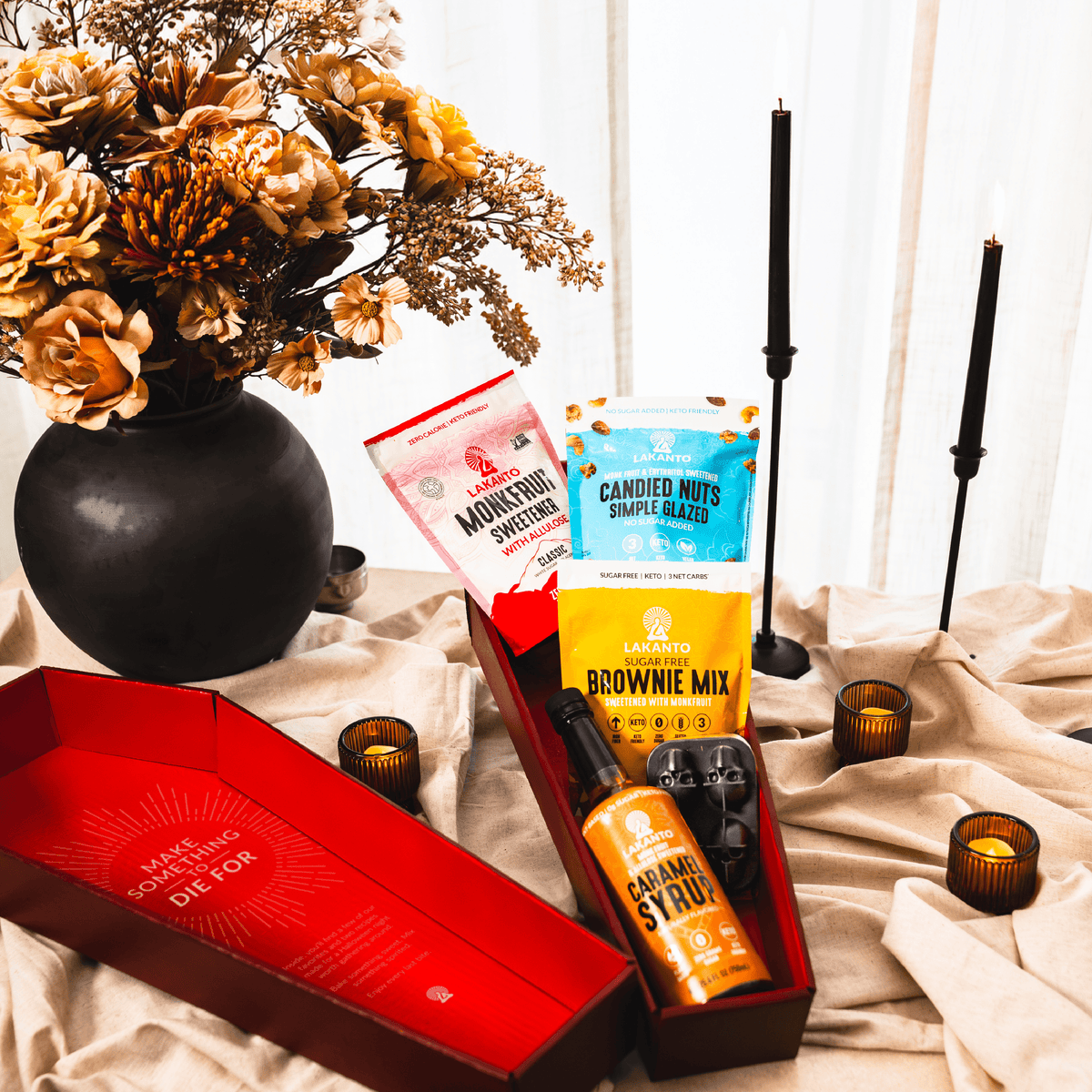 A styled gift set arranged on draped beige fabric with black candles, a black vase of dried flowers, and amber candleholders. The set includes an open red box labeled 'MADE WITH A SPOON BY DILOR,' a brownie mix bag, a candy corn syrup bottle, a mountain dew sweetener packet, and a bag of candied nuts.