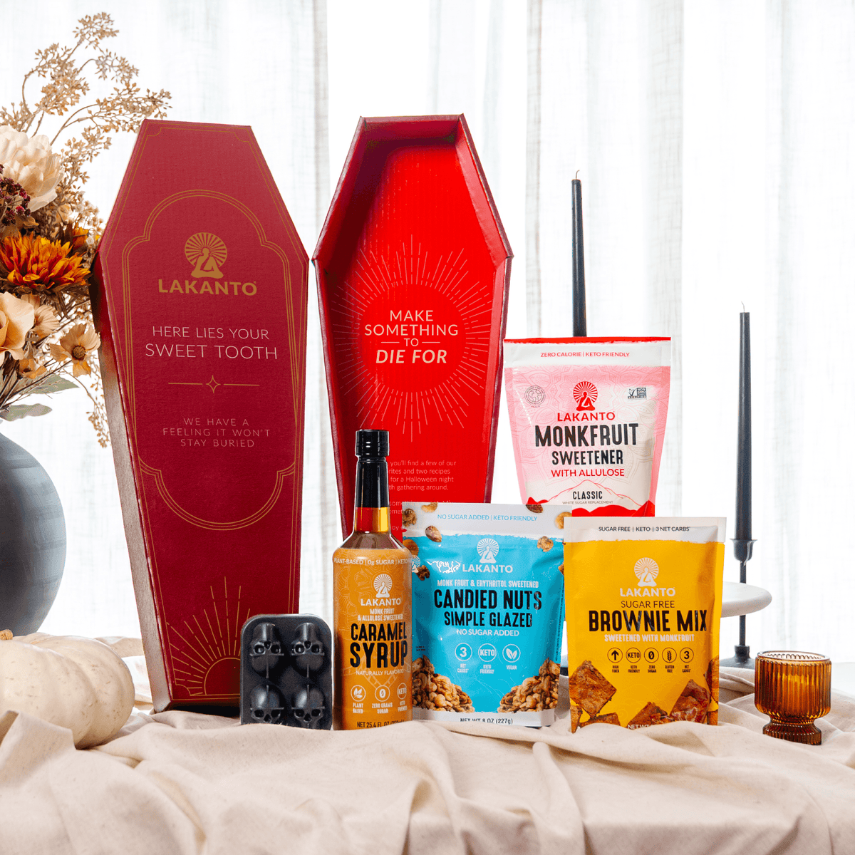 A curated snack and candy gift set arranged on a draped cream fabric surface. The set includes two red coffin-shaped Lakanto gift boxes, a bottle of caramel syrup, a bag of caramel corn, a bag of candied nuts, a bag of brownie mix, and a bag of Monkbfruit sweetener, with decorative flowers and candles in the background.