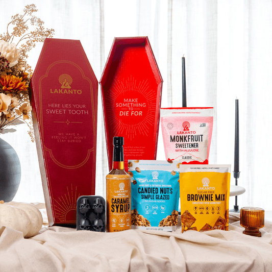 A curated snack and candy gift set arranged on a draped cream fabric surface. The set includes two red coffin-shaped Lakanto gift boxes, a bottle of caramel syrup, a bag of caramel corn, a bag of candied nuts, a bag of brownie mix, and a bag of Monkbfruit sweetener, with decorative flowers and candles in the background.