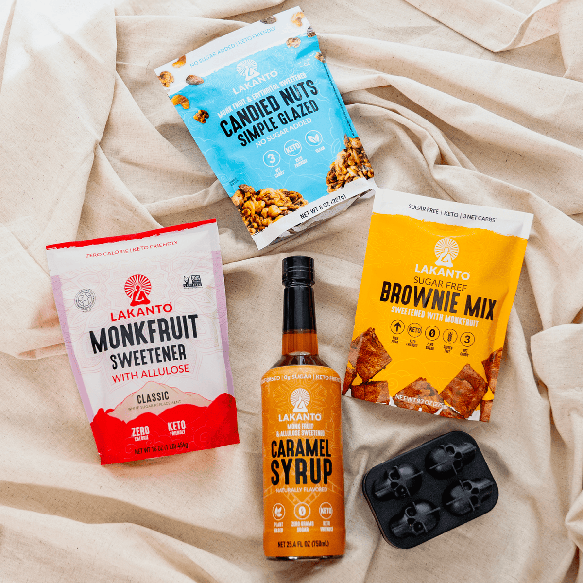 A flat-lay arrangement of Lakanto sugar-free snack products on a beige fabric background, including a bottle of caramel syrup, a bag of monkfruit sweetener, a bag of caramelized nuts, a bag of brownie mix, and a black ice mold tray.
