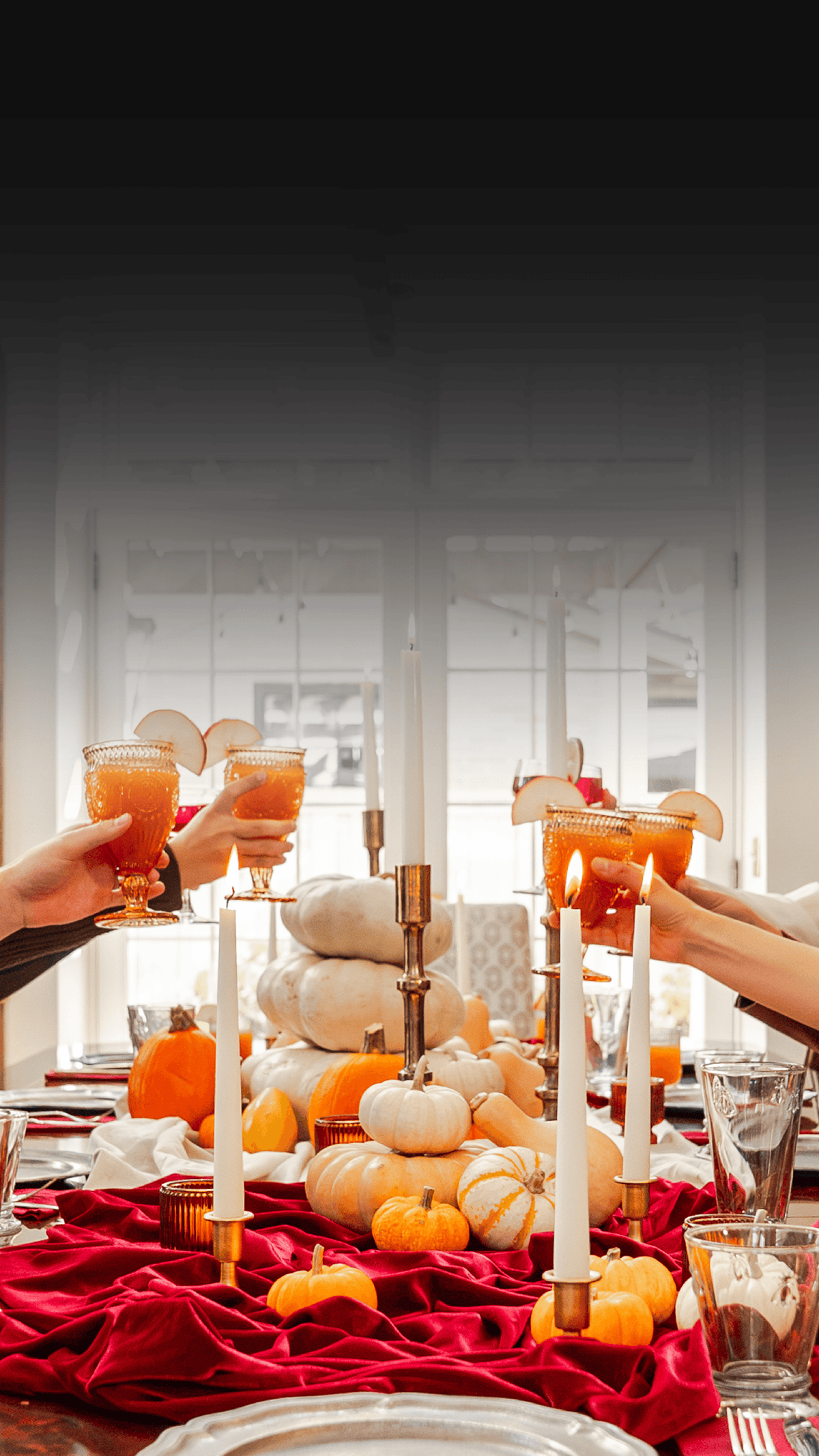 A festive autumn table setting with a red draped tablecloth, white and orange pumpkins, lit taper candles in gold holders, and multiple hands holding orange cocktails in stemmed glasses, set against a softly blurred bright window background.
