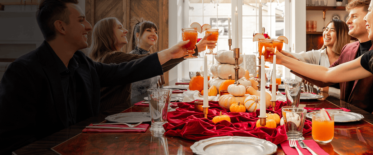 A group of people raising orange cocktails in a toast at a decorated dining table with candles, pumpkins, and a red table runner in a warm indoor setting.