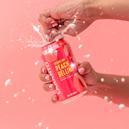 Hand holding and opening a pink can of Lakanto Sparkling Peach Bellini Mocktail drink against a pink background. Naturally flavored. Monk fruit with allulose sweetened.