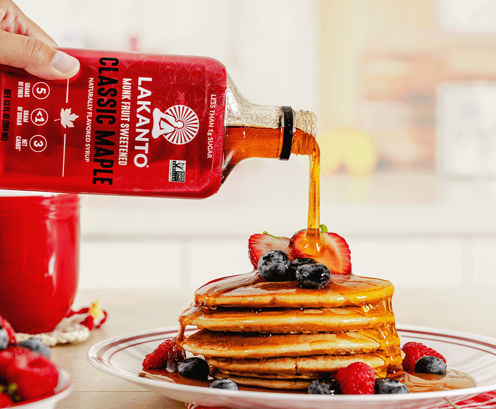 A hand pours a bottle of Lakanto Classic Maple syrup over a stack of pancakes topped with strawberries and blueberries on a plate, with a red mug in the blurred background.